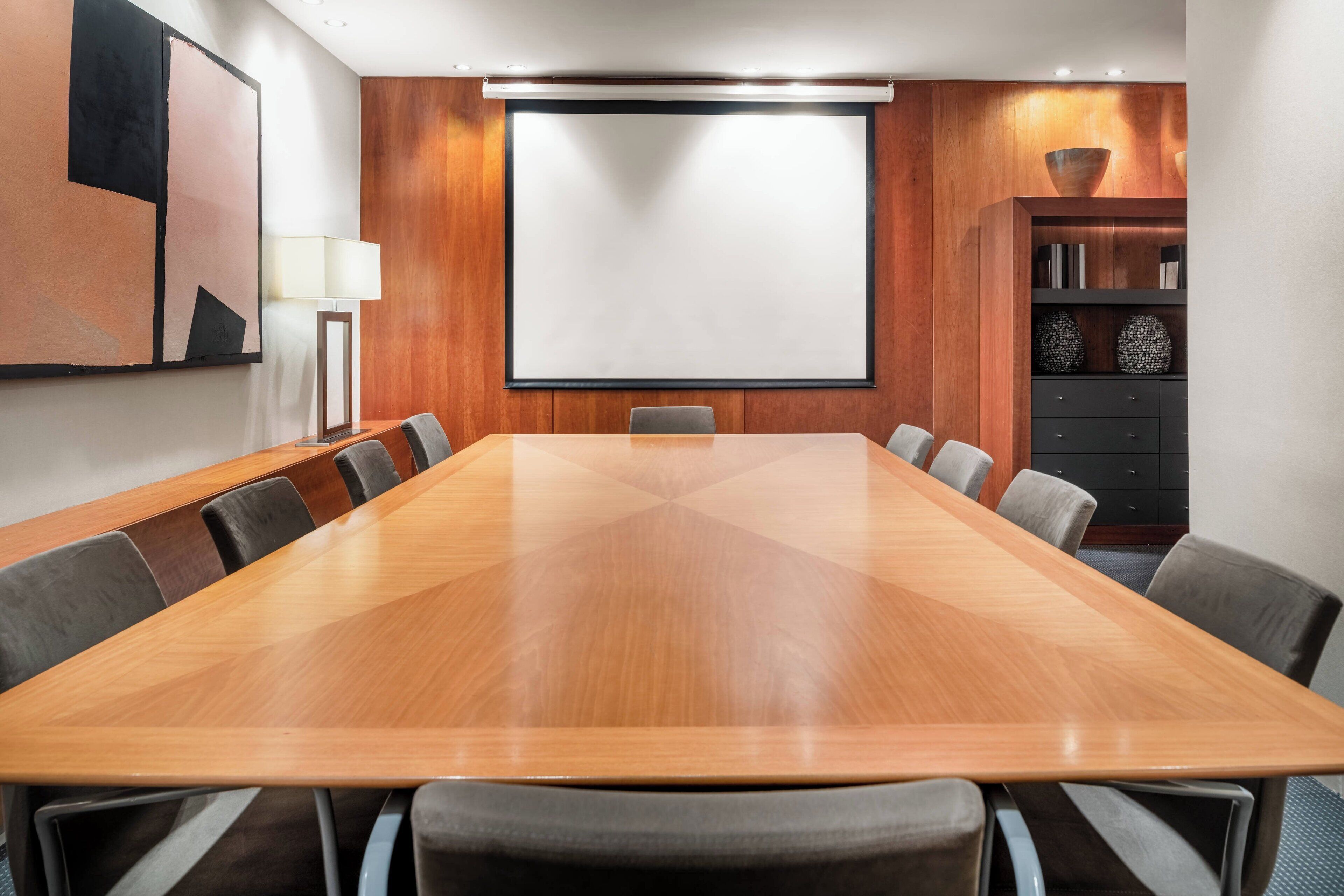 A conference room with a large wooden table, grey chairs, a projector screen, and a cabinet with decorative items.