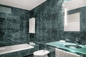 A modern bathroom with a bathtub, a sink, and a bidet. The walls are covered in dark marble tiles. - AC Hotel Badajoz by Marriott (Badajoz)