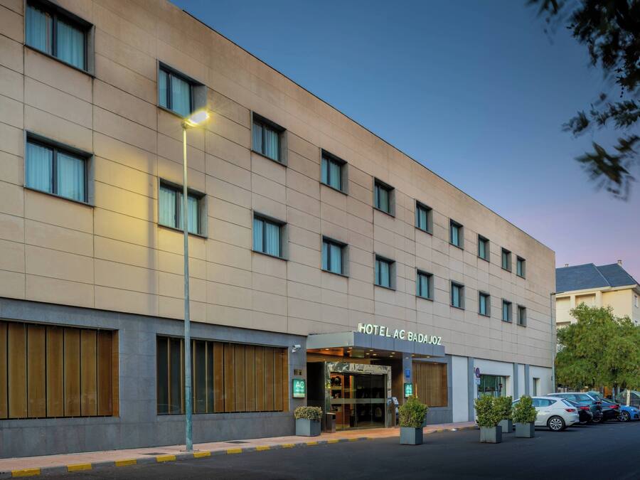 Een hotel met een moderne gevel, een goed verlichte entree en een parkeerplaats met verschillende auto's.