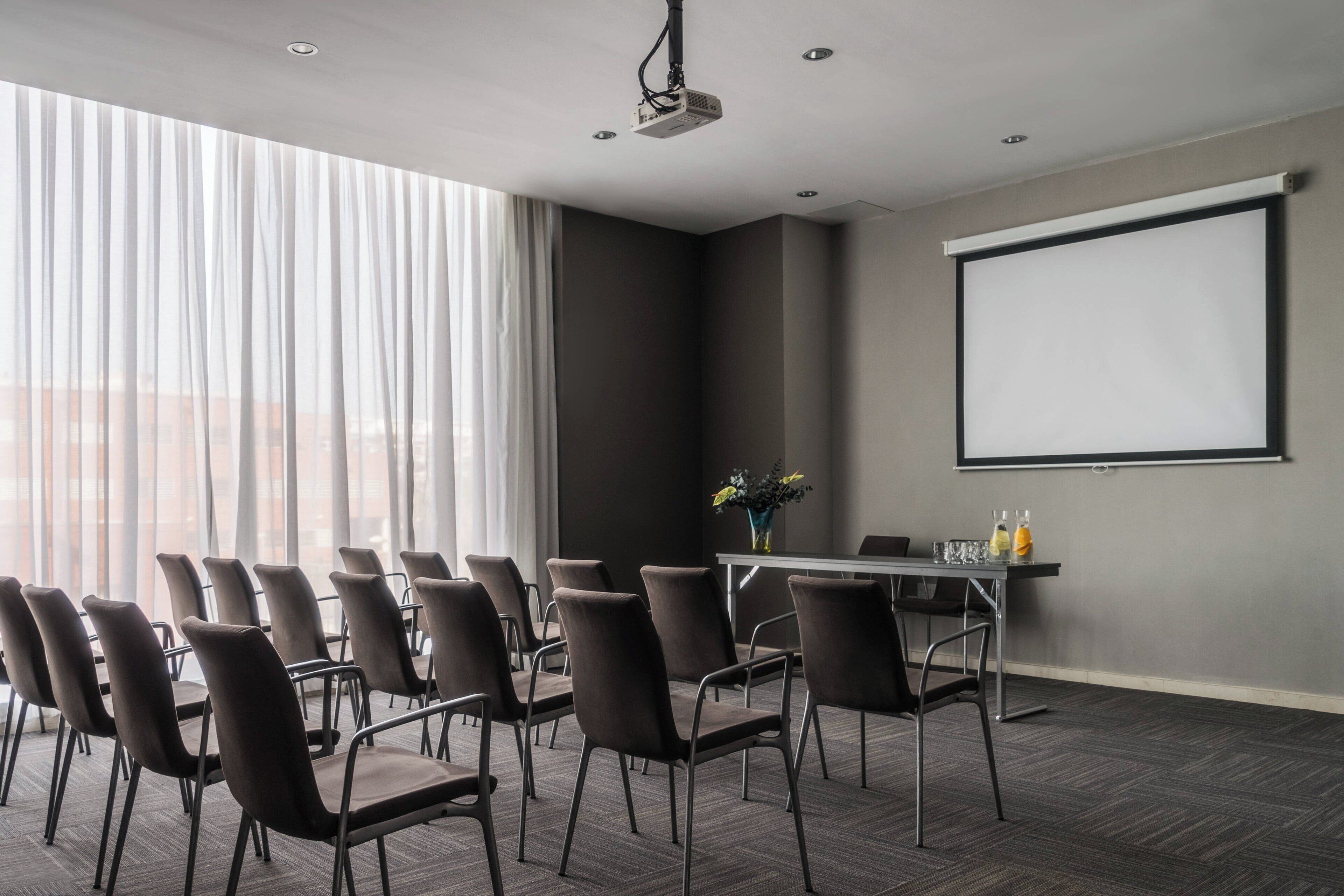 Uma sala de conferências com projetor, uma tela grande e uma mesa com cadeiras.