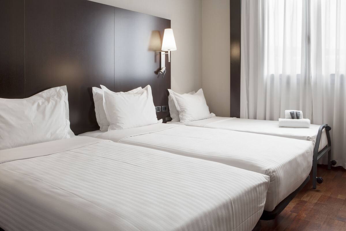 A hotel room with two beds, a dark headboard, a wall-mounted lamp, and a window with sheer curtains.