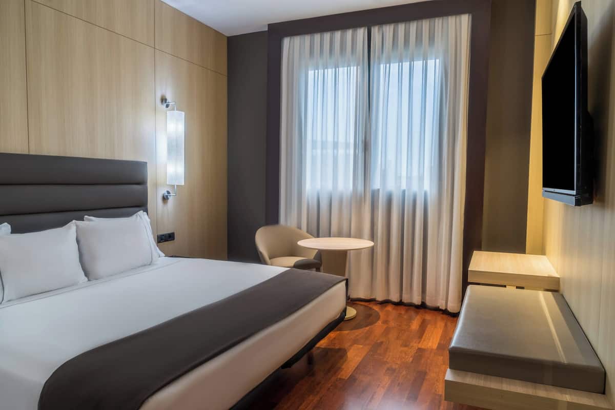 A modern hotel room with a large bed, a small round table, a chair, and a TV mounted on the wall.