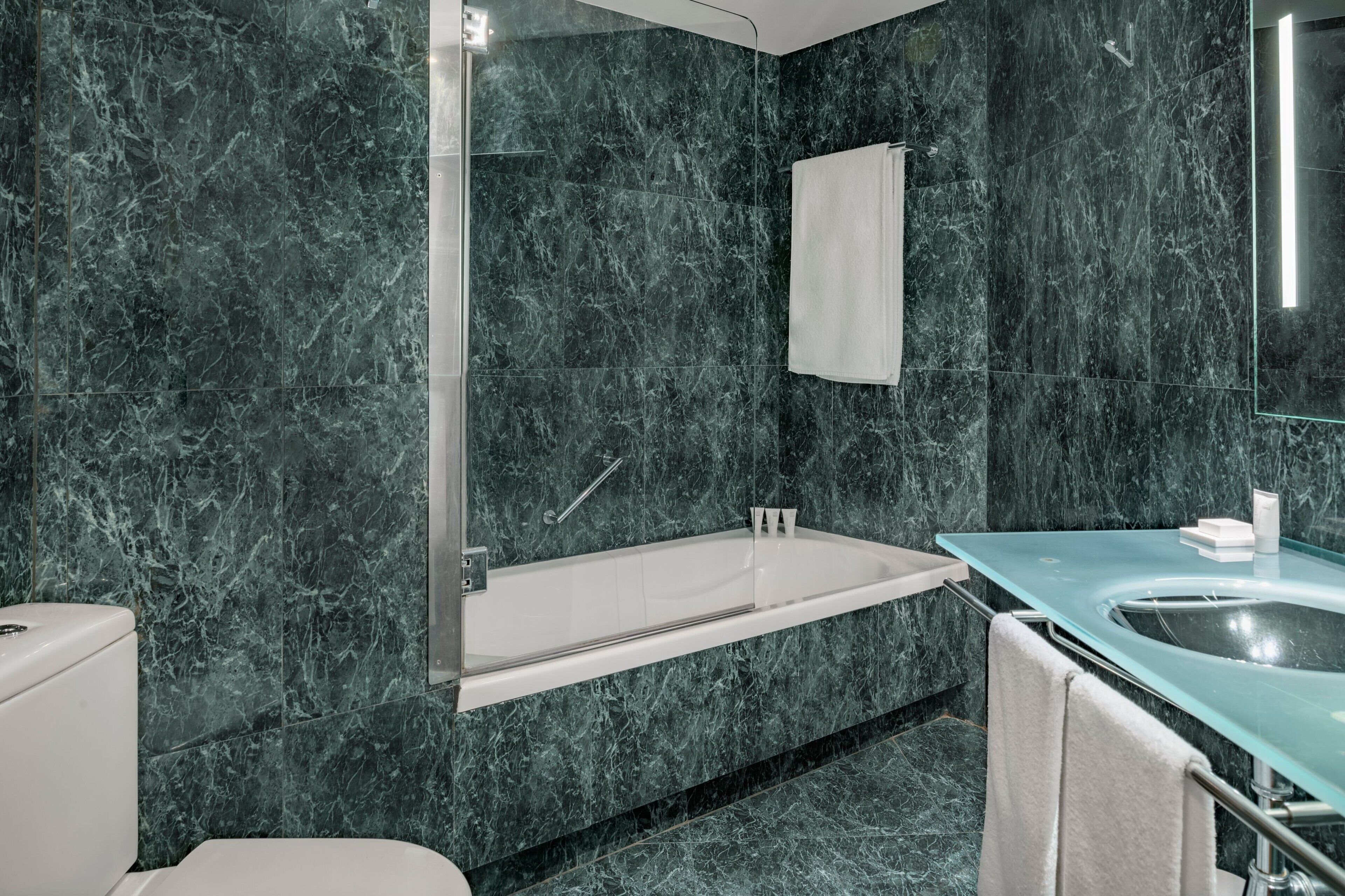 Un baño moderno con bañera de hidromasaje envuelta en vidrio, inodoro blanco y lavamanos con espejo.