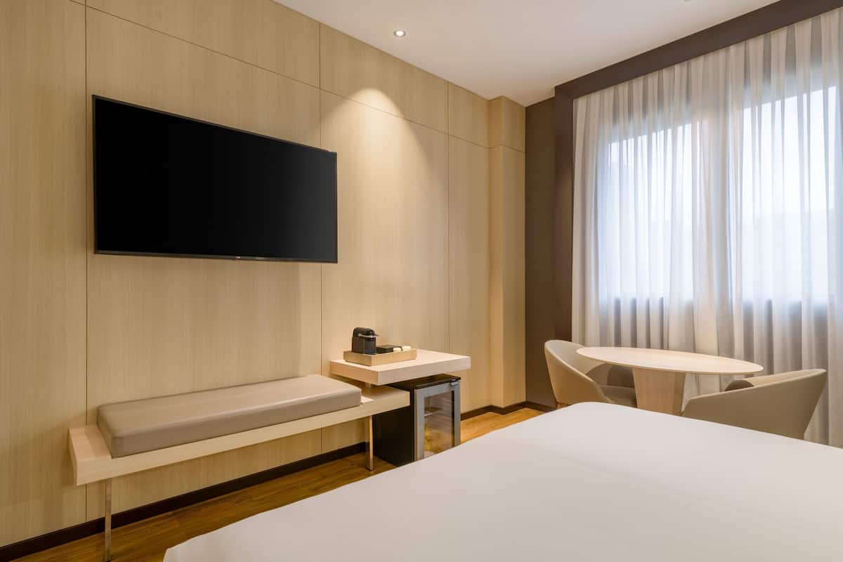 A modern hotel room with a large bed, a flat-screen TV mounted on the wall, a small table with chairs, and a window with curtains.