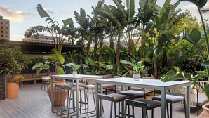 Une terrasse extérieure dotée d’une longue table et de plusieurs tabourets de bar, entourée d’une végétation tropicale luxuriante et d’une plateforme en bois.
