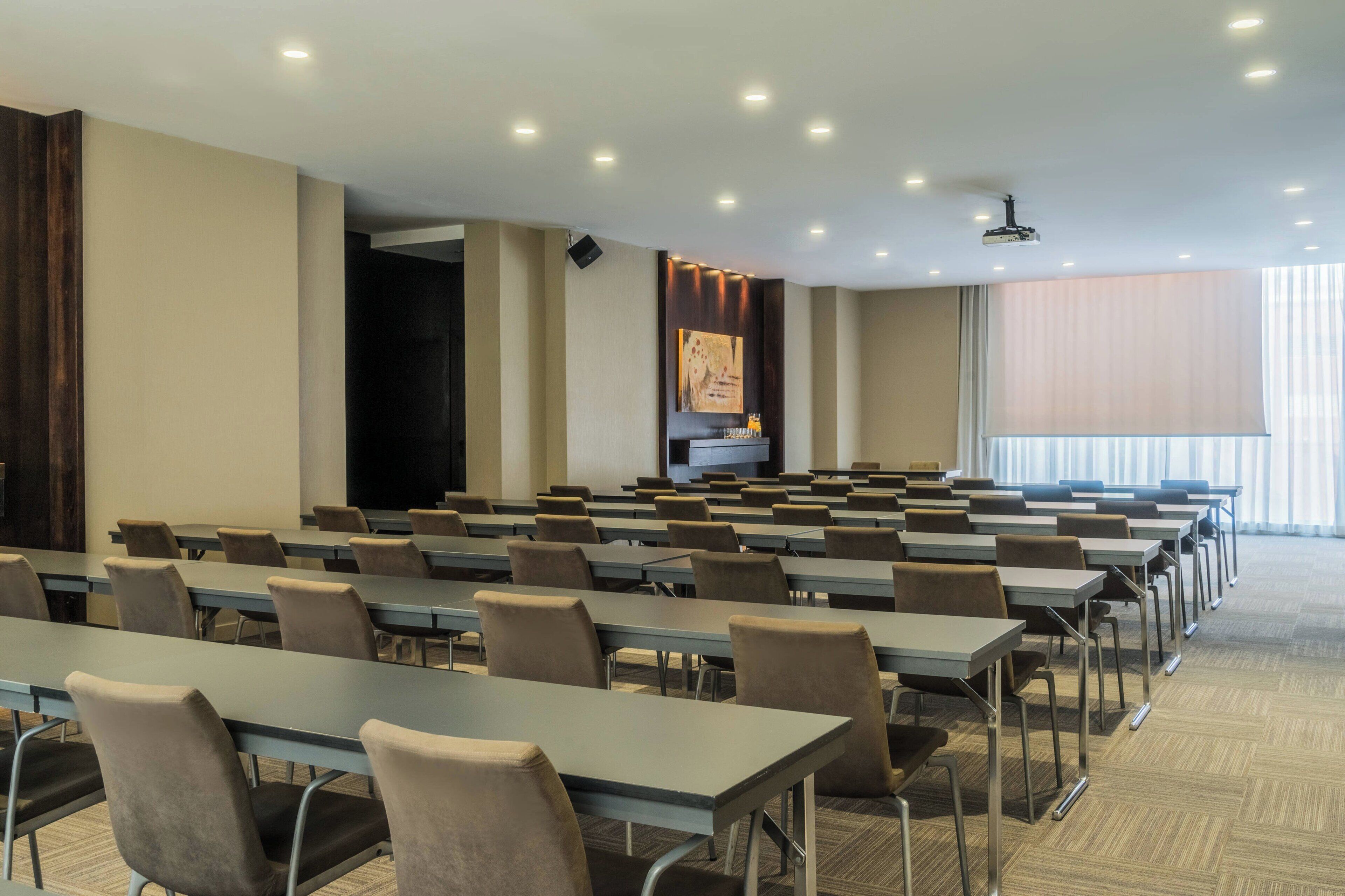 Un salón de conferencias con filas de mesas y sillas, una pantalla de proyector y un amplio ventanal con cortinas.