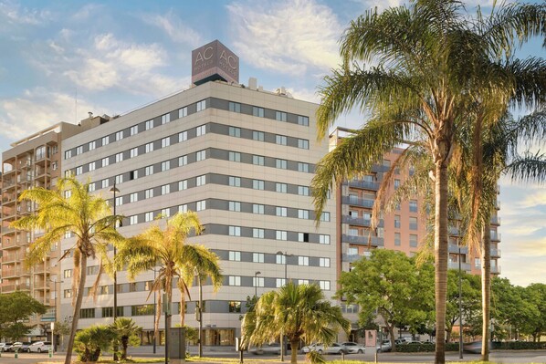Un hotel de varios pisos con un diseño arquitectónico moderno, que presenta un llamativo logotipo de AC Hotels en la azotea.