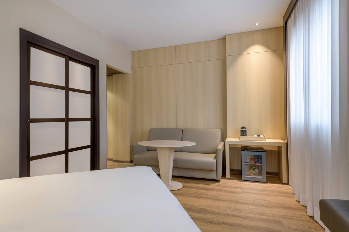 A hotel room with a bed, a round table, a sofa, a small kitchenette, and a sliding door.