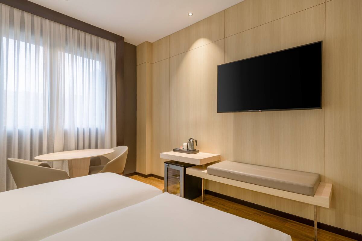 A hotel room with two beds, a TV mounted on the wall, a small table with a kettle, and a window with curtains.