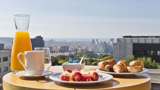 A breakfast setup with a glass of orange juice, a white mug, a plate of strawberries and watermelon, a bowl of yogurt with a spoon, and a plate of croissants, all on a table with a city view in the background. - Abba Garden (Esplugues de Llobregat)