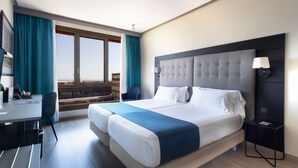 A hotel room with a large bed, a desk, and a chair. There is a window with curtains, a lamp, and a view of buildings outside. - Abba Garden (Esplugues de Llobregat)