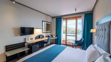 Habitación de hotel con cama, escritorio, silla, televisor y balcón con cortinas.