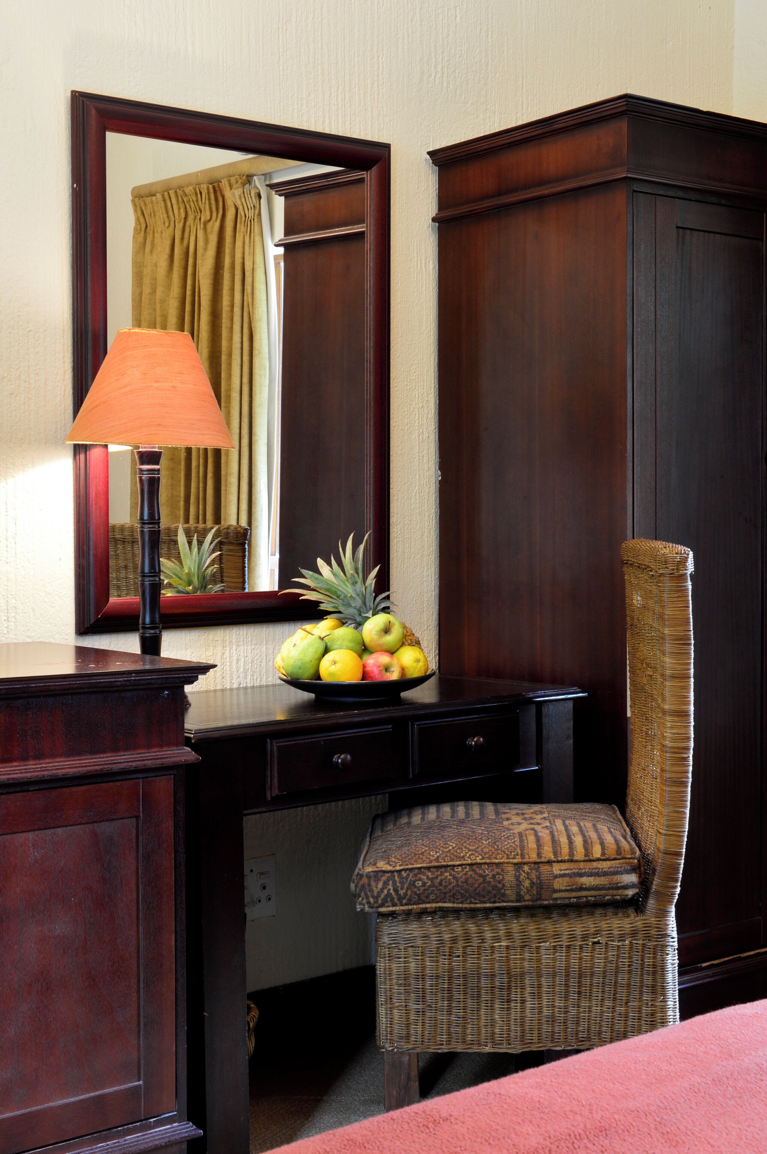 A hotel room with a wooden desk, a lamp, a chair, a mirror, and a fruit bowl.