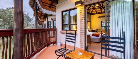 A balcony with a wooden table and chairs, a brick wall, and a glass door leading to a room with a sofa and a lamp.