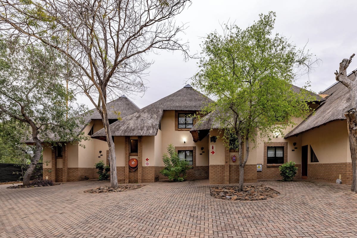 A building with multiple rooms, each with a thatched roof and brick walls. There are trees in front of the building, and a paved area in front.