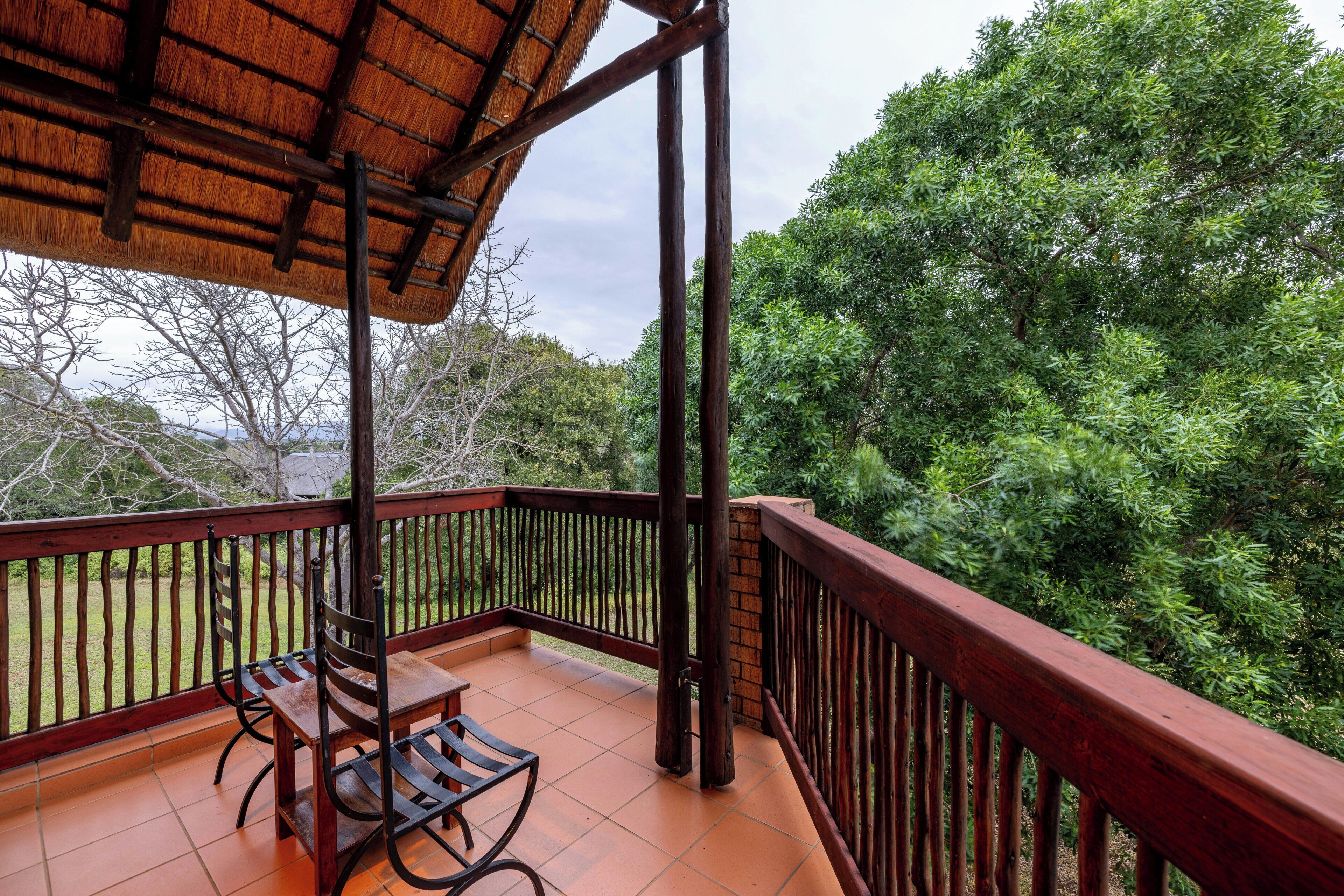 A covered outdoor area with a wooden table and chairs, surrounded by a wooden railing, overlooking a lush green landscape.