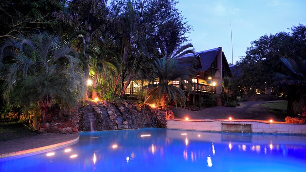 A swimming pool with a stone wall and palm trees, surrounded by a building with a balcony. - Pestana Kruger Lodge (Nkomazi)