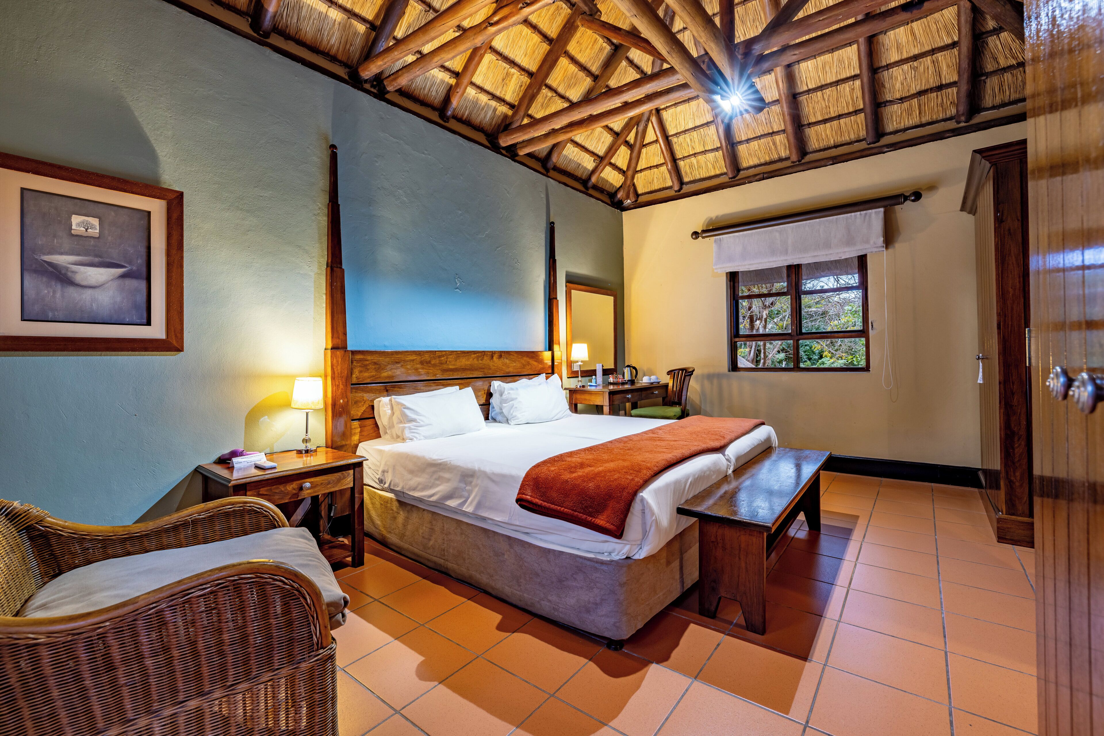 A hotel room with a bed, a bench, wicker furniture, a wooden ceiling, a window, and a framed picture on the wall.