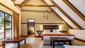 A bedroom with a sloped ceiling, a bed, a bench, a bedside table, and a window with curtains. - Pestana Kruger Lodge (Nkomazi)