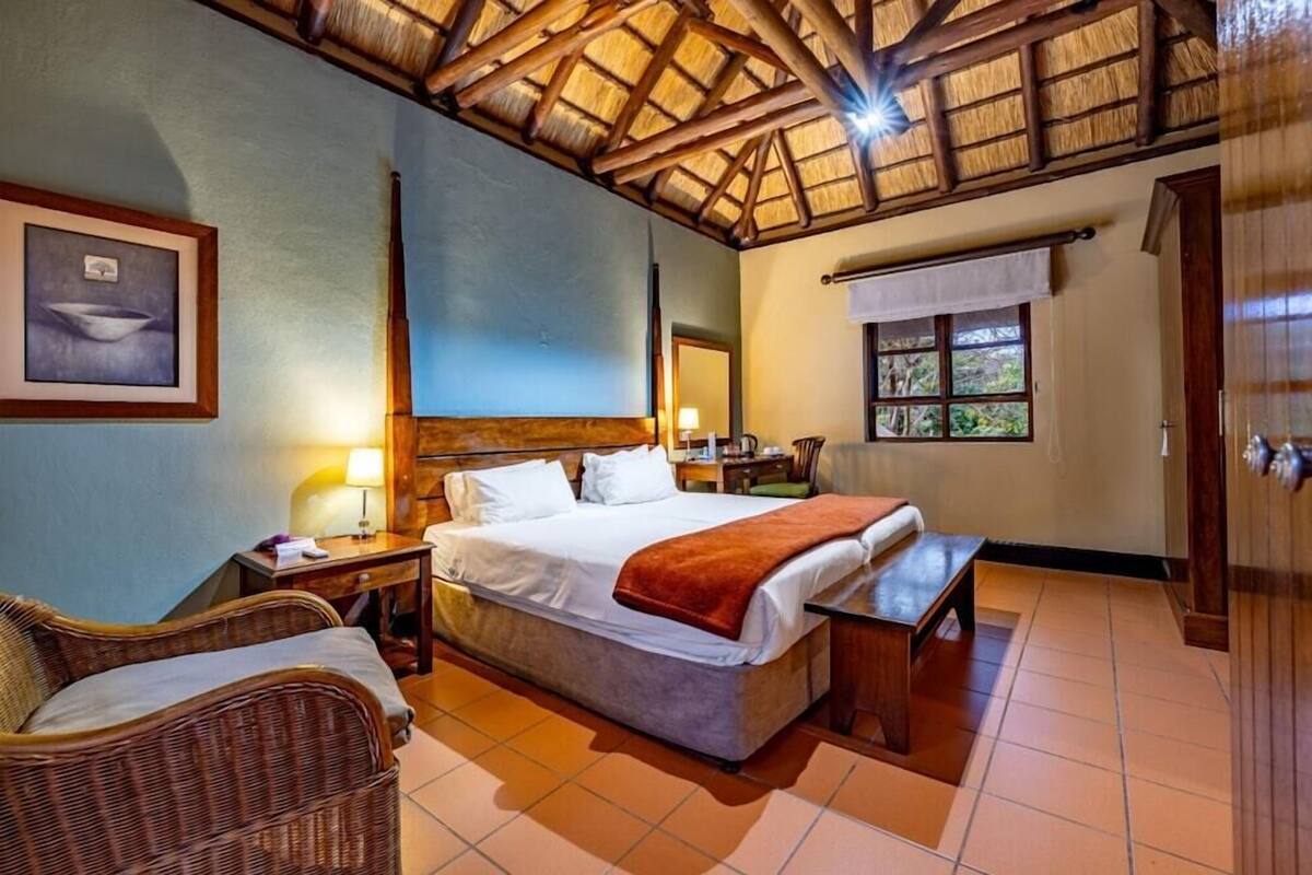 A room with a bed, a wicker chair, a desk, a window, and a wooden ceiling.
