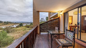 A balcony with a wooden floor, a glass table, a chair, and a view of a river and mountains.