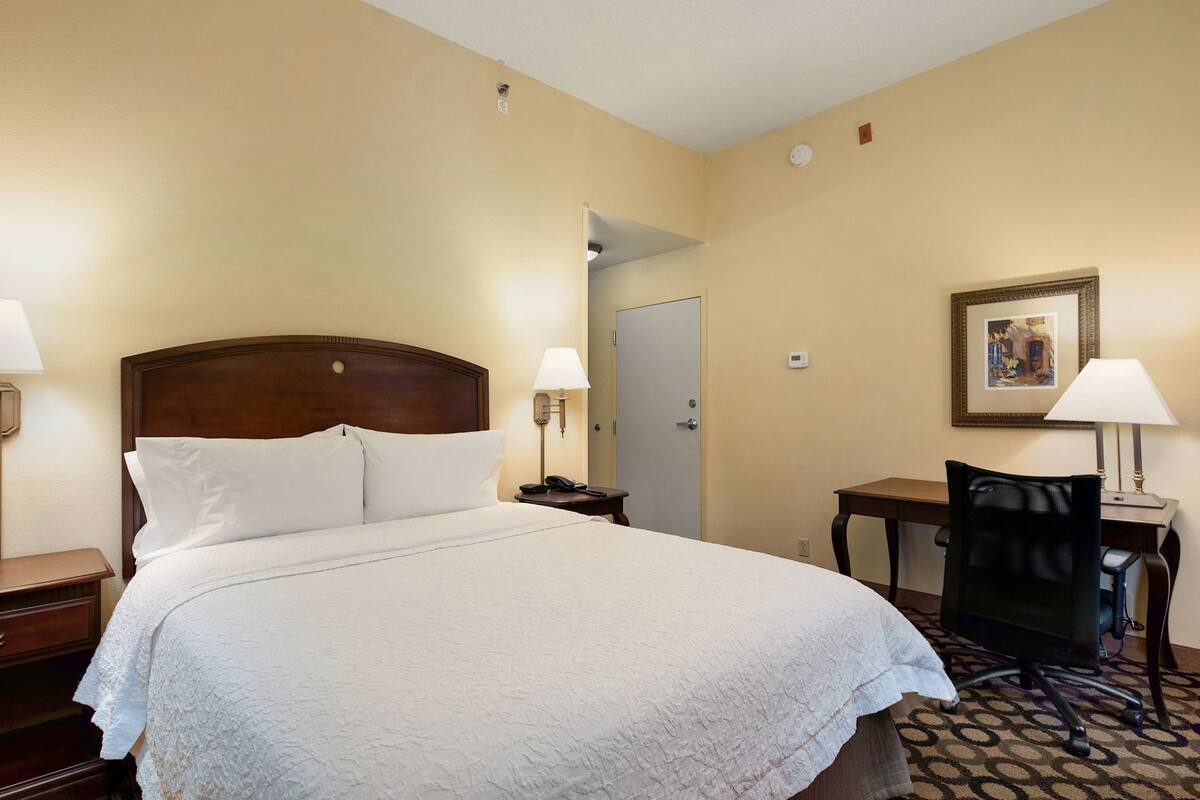 A hotel room with a large bed, bedside lamps, a desk, a chair, and a door.