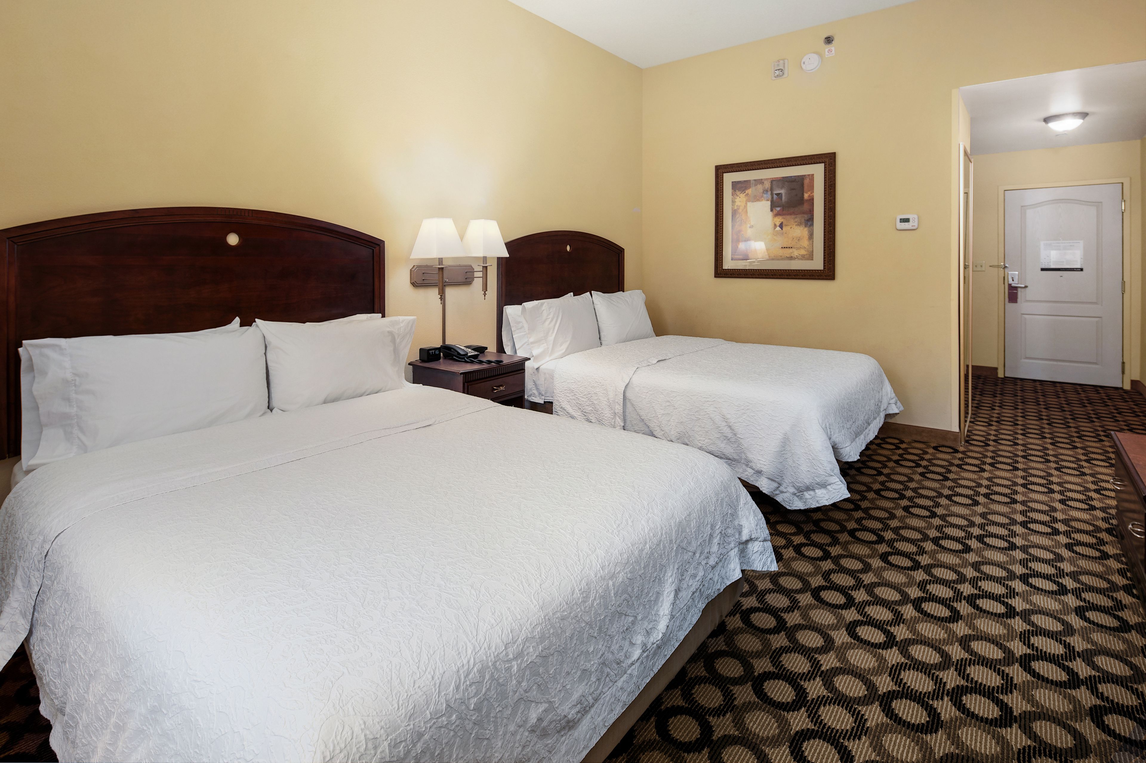 A hotel room with two beds, a nightstand with a lamp, a framed picture on the wall, and a door with a lock.