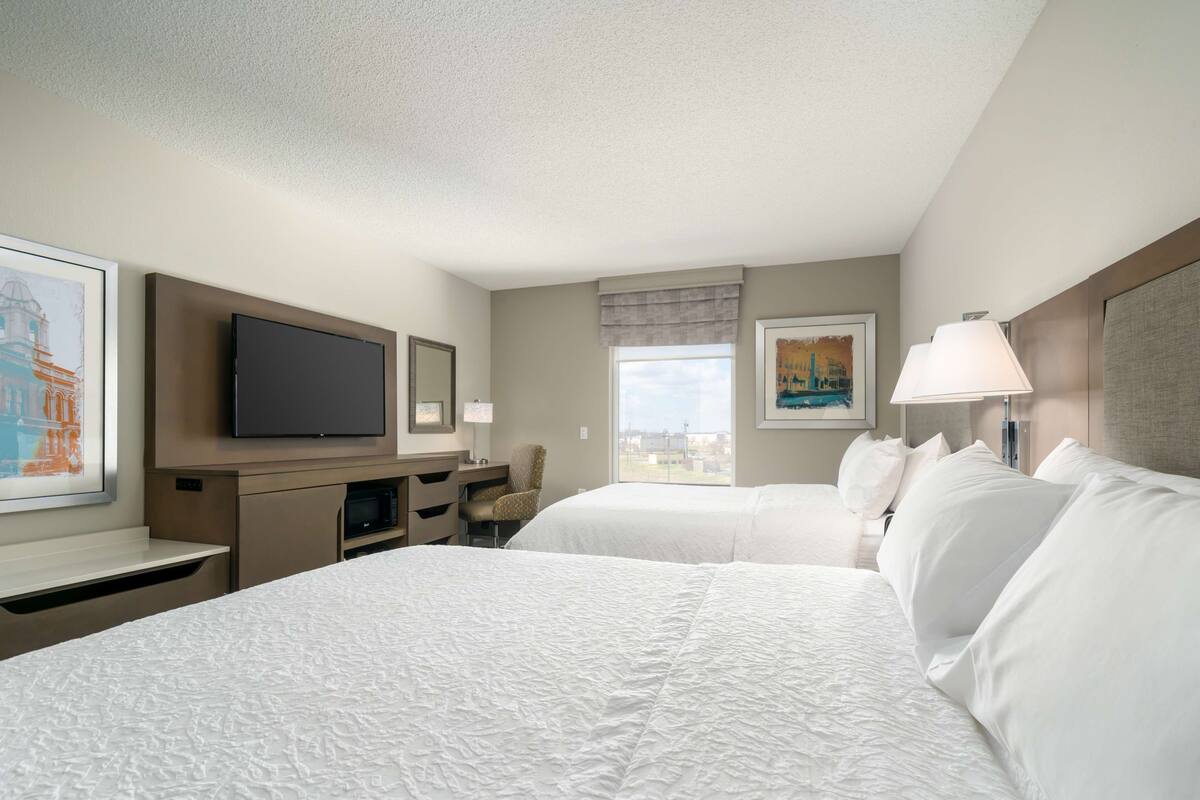 A hotel room with two beds, a flat-screen TV, a desk, and a window with a city view.