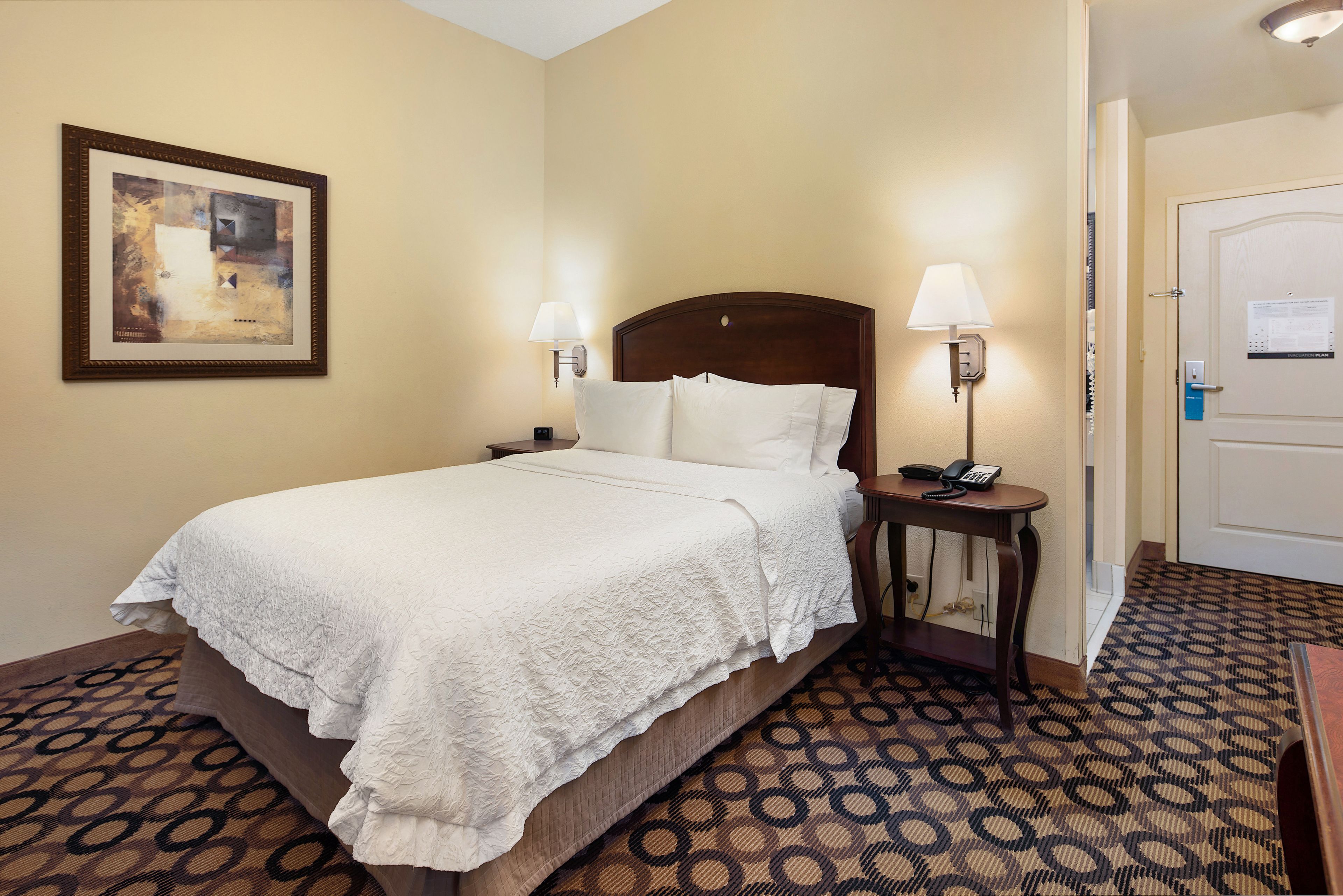 A hotel room with a large bed, two bedside tables with lamps, a framed artwork on the wall, and a patterned carpet.