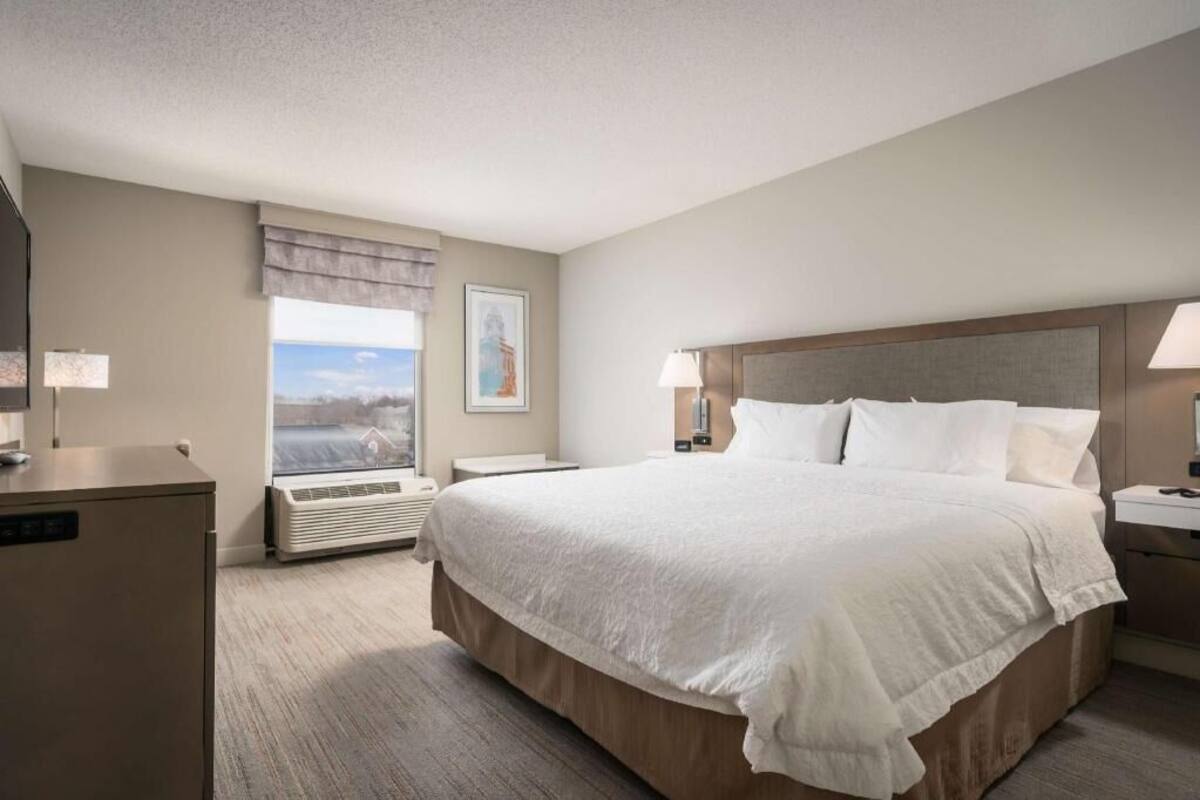 A hotel room with a large bed, a desk, a chair, a window with a view, and a wall-mounted air conditioner.