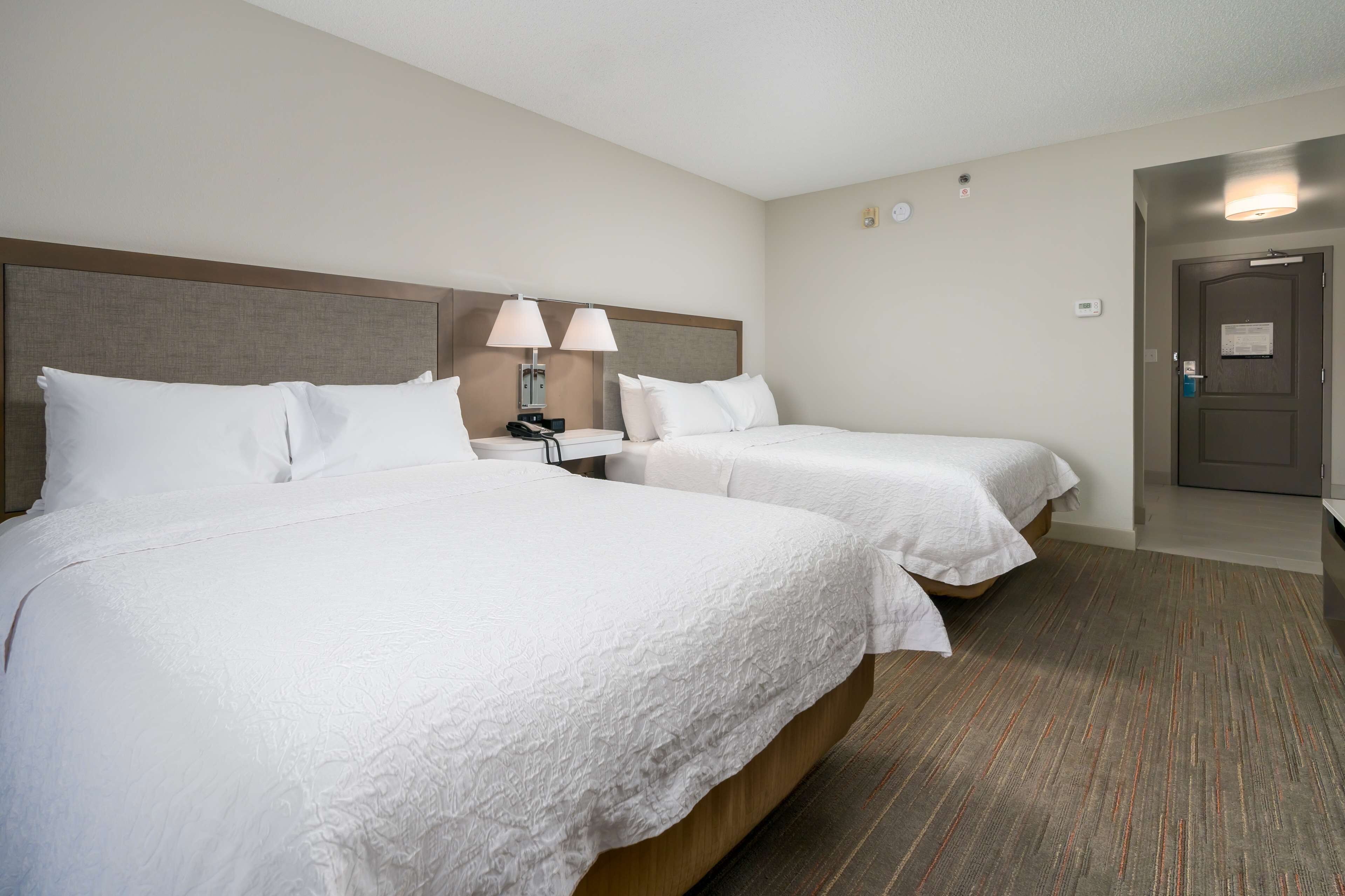 A hotel room with two beds, a nightstand with a lamp, a wall clock, and a door with a handle.