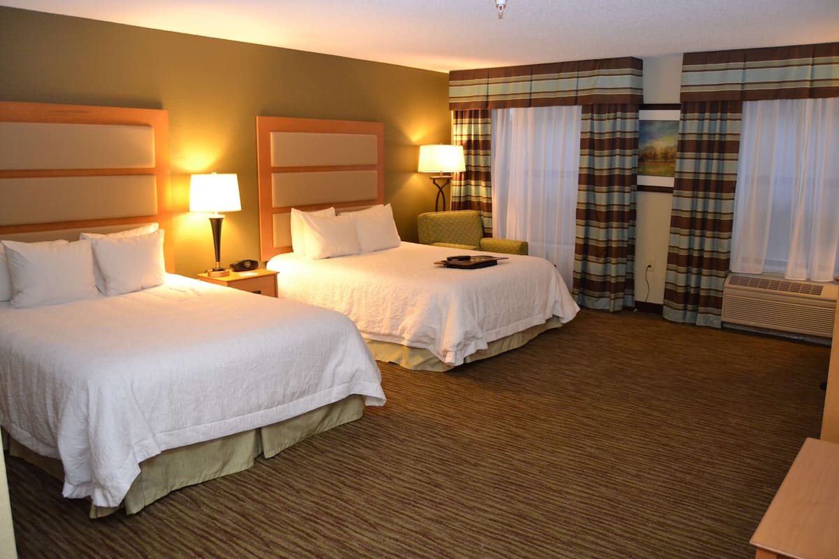 Habitación de hotel con dos camas, una mesita de noche con lámpara, una silla y una ventana con cortinas.