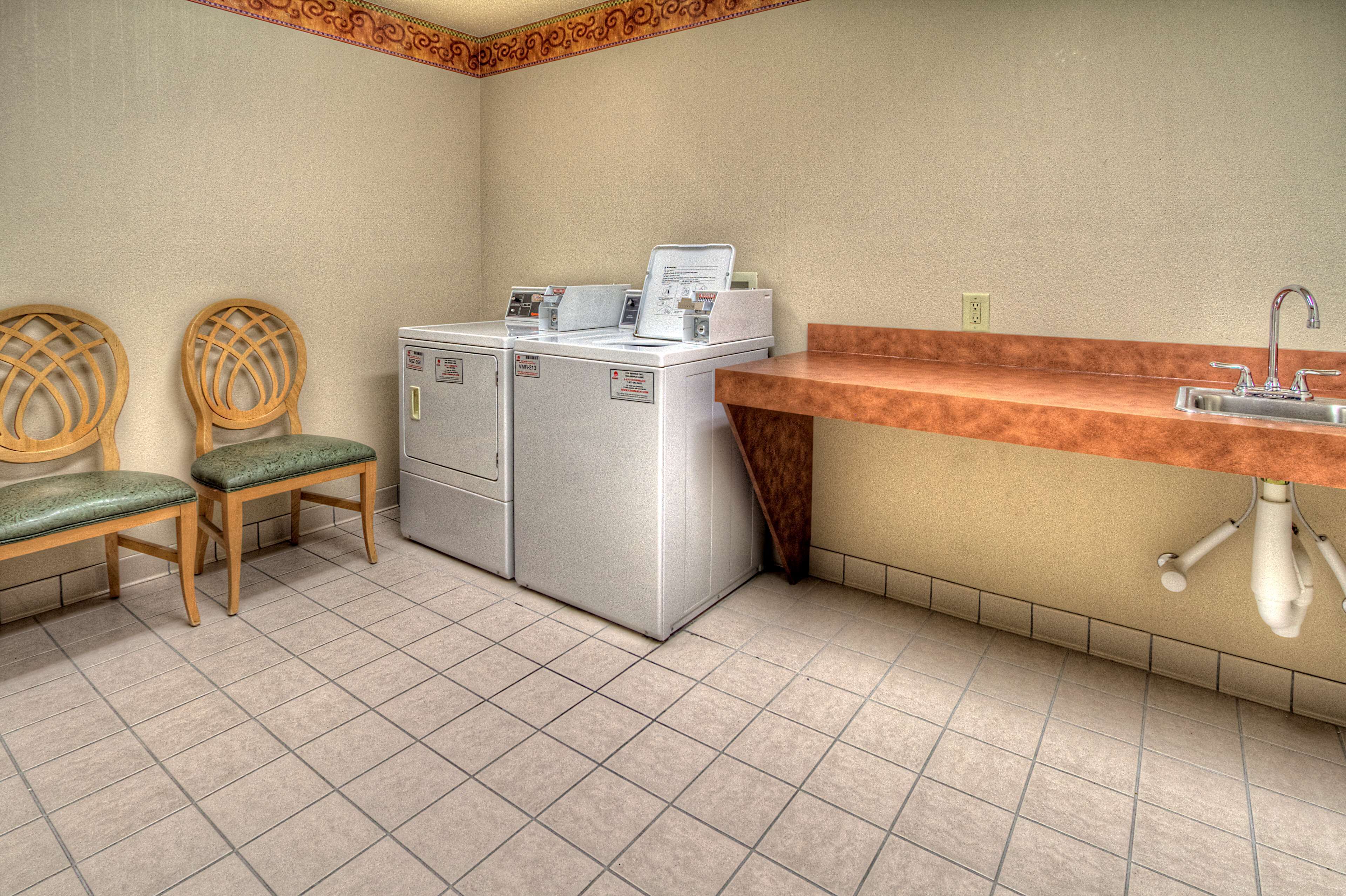 A laundry area with two washing machines, a sink, and two wooden chairs with green cushions.