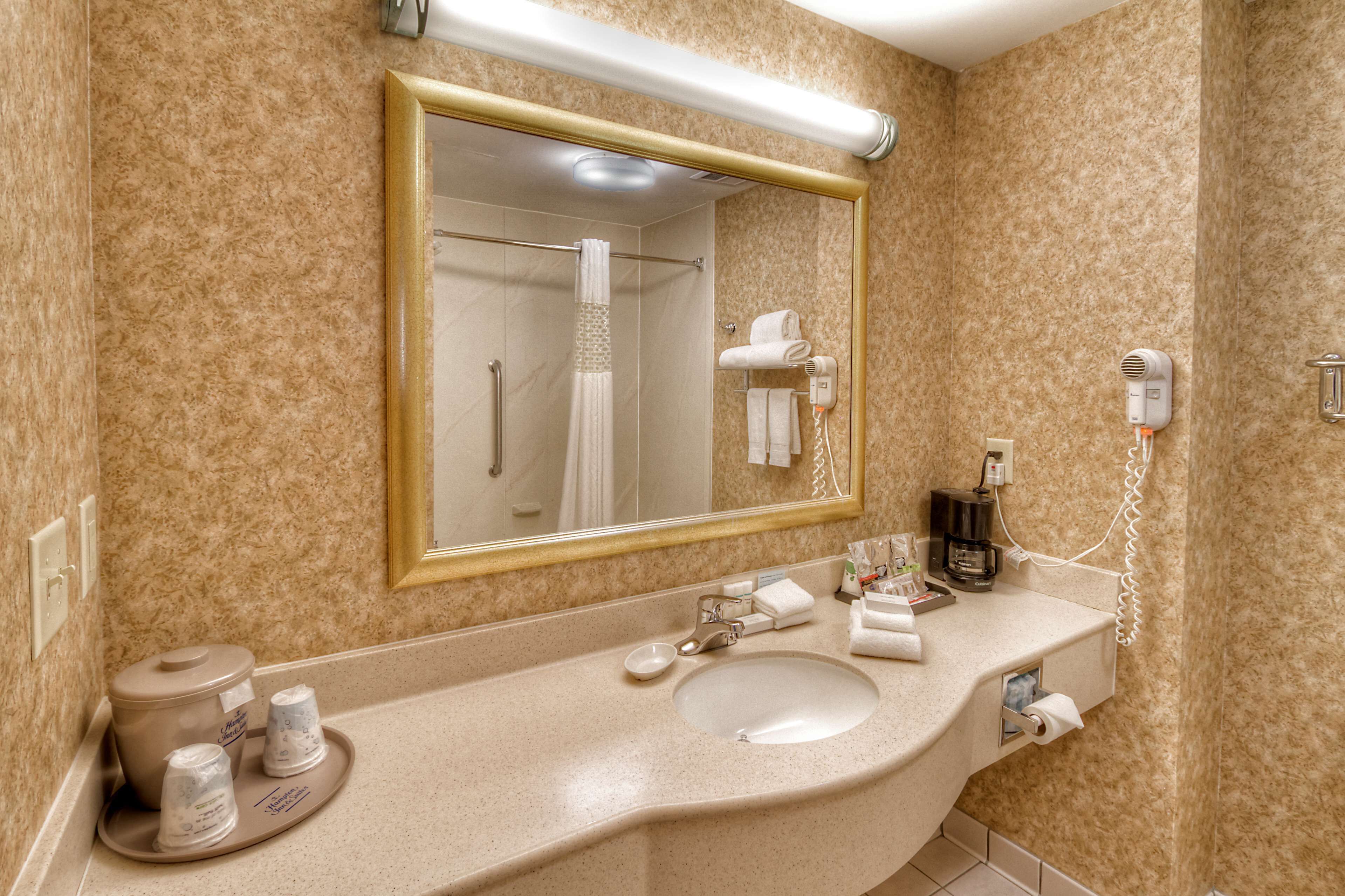 Casa de banho de hotel com um espelho grande, uma pia, um porta-toalhas, um secador de cabelo e uma máquina de café.