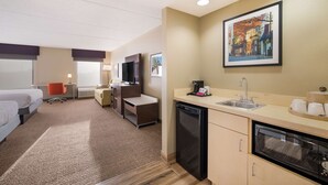 A hotel room with a bed, a desk, a chair, a small kitchenette with a sink and a dishwasher, and a painting on the wall.
