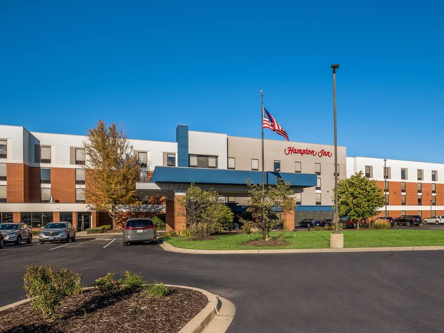 A modern hotel with a flat roof, a flagpole with an American flag, and a parking lot with several cars.