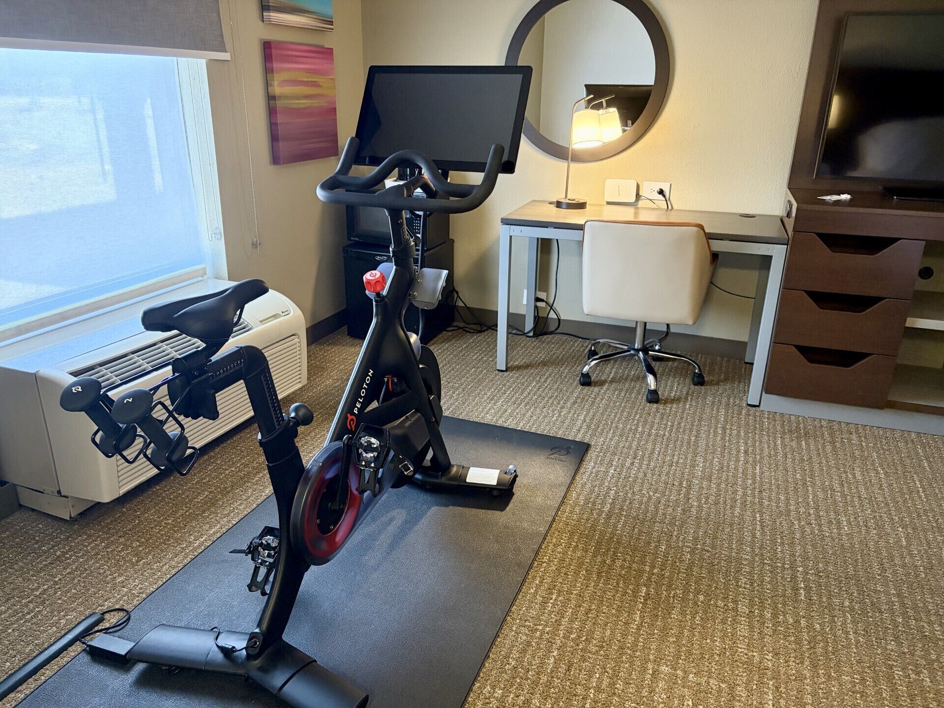 An office space with a stationary bike, a desk with a computer, a chair, and a large mirror.