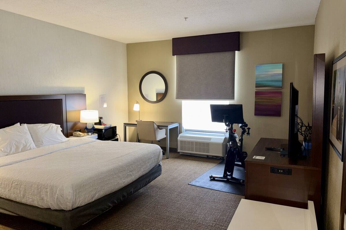 Een hotelkamer met een bed, bureau, stoel, lamp, spiegel, kunstwerk en een televisie.