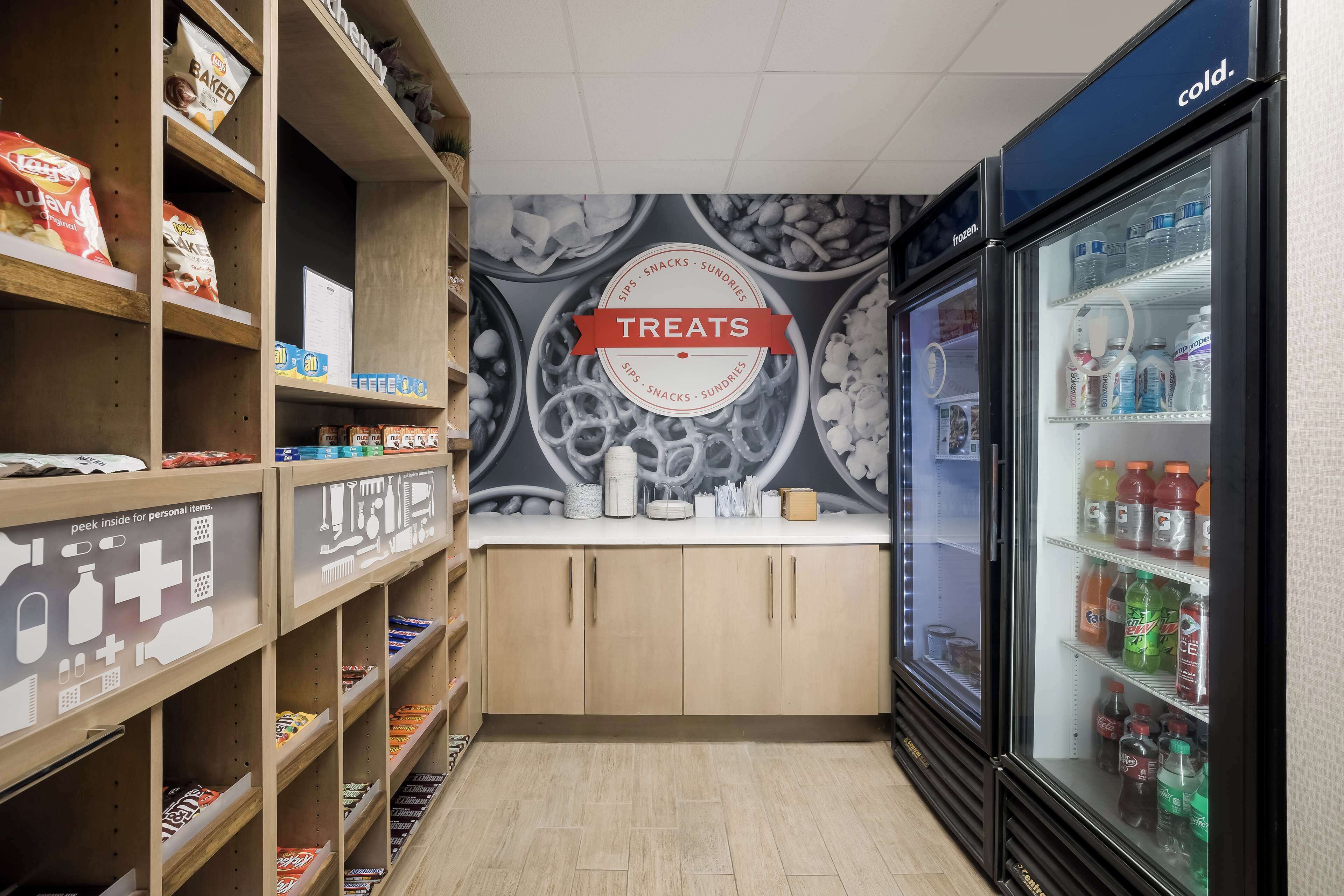 Un magasin dont les étagères sont remplies d’en-cas et de friandises, un rayon réfrigéré avec des boissons, et un mur orné d’une enseigne indiquant « FRIANDISES ».