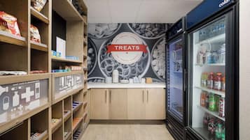 Uma loja com prateleiras repletas de lanches e guloseimas, uma área refrigerada com bebidas e um painel com a inscrição 'TREATS'.