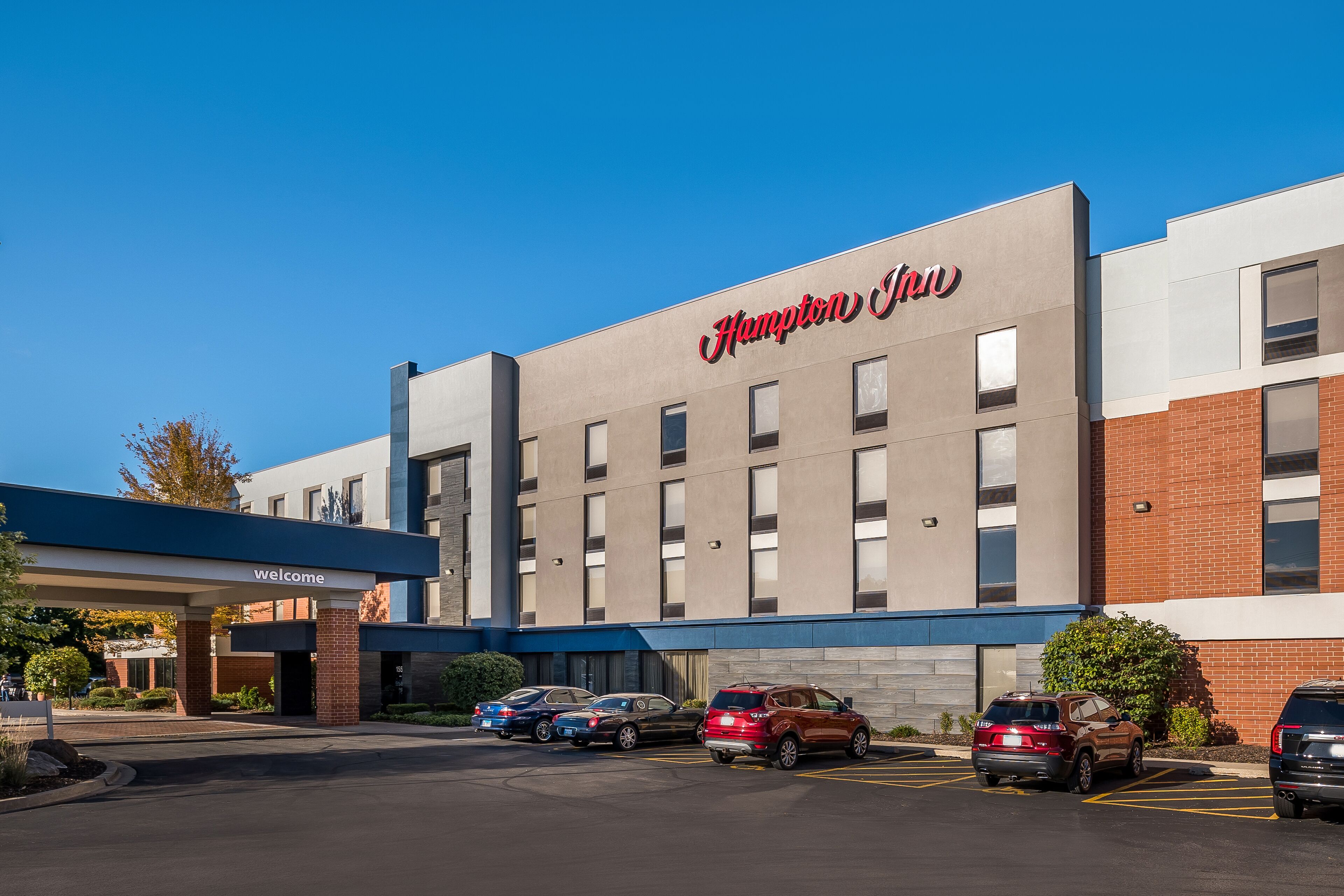 Modernilla julkisivulla varustettu Hampton Inn -hotelli, useita autoja sisältävä parkkipaikka ja kirkkaan sininen taivas.