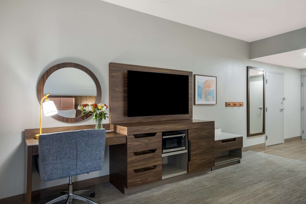 A modern hotel room with a wooden entertainment unit, a desk with a chair, a large mirror, and a microwave.