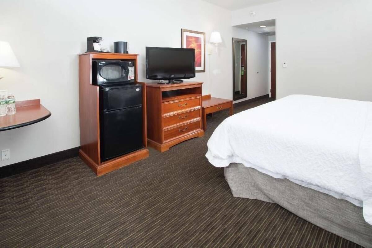 A hotel room with a bed, a television, a minibar, and a desk with a lamp.