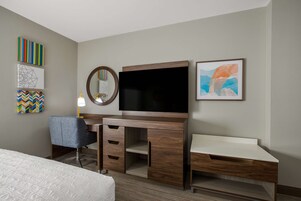 A hotel room with a bed, a desk with drawers, a television, a mirror, and artwork on the walls.
