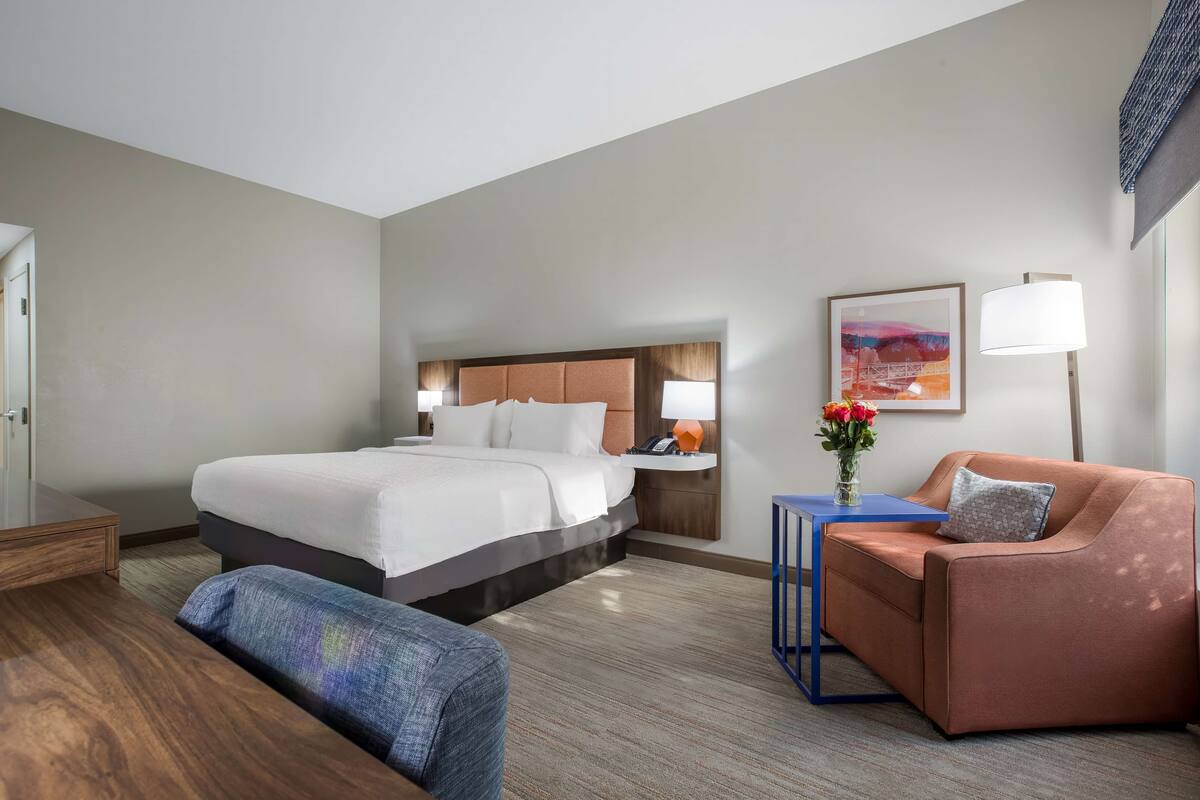 A hotel room with a large bed, a desk, a chair, a sofa, and a nightstand with a lamp.