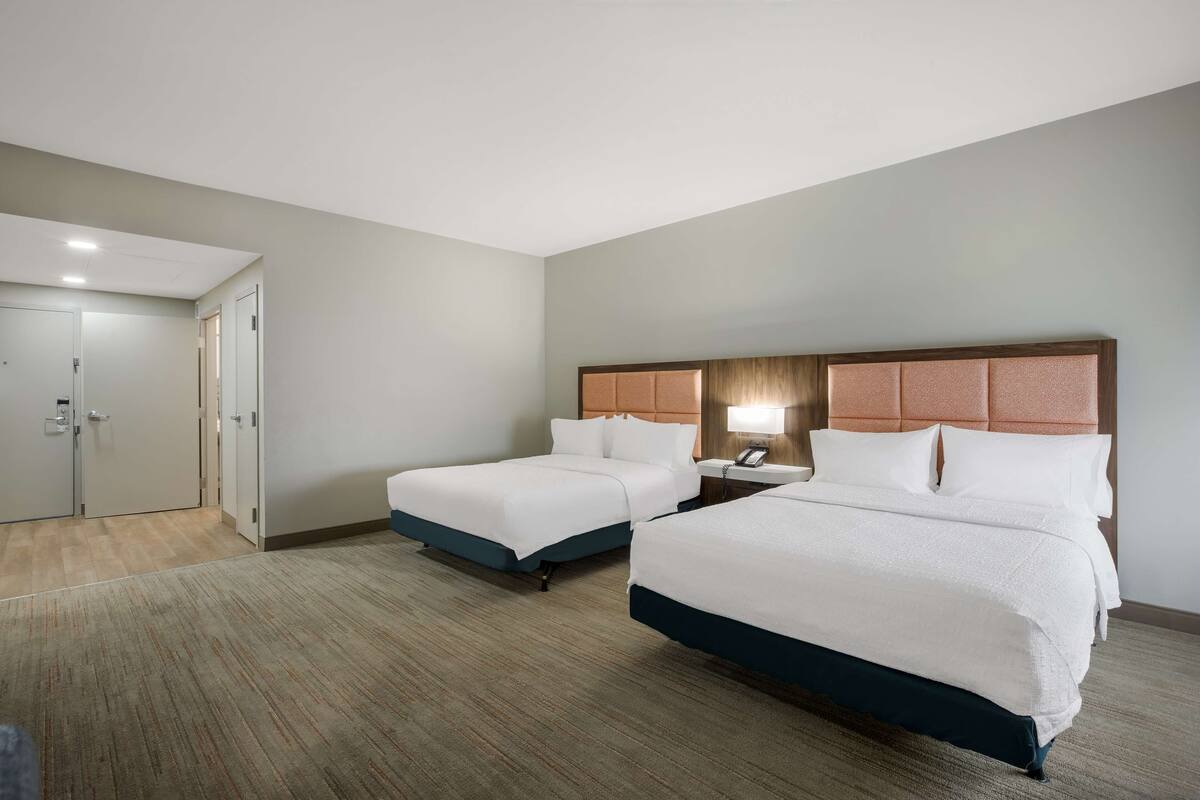 A hotel room with two beds, a wooden headboard, a bedside table with a lamp, and a carpeted floor.