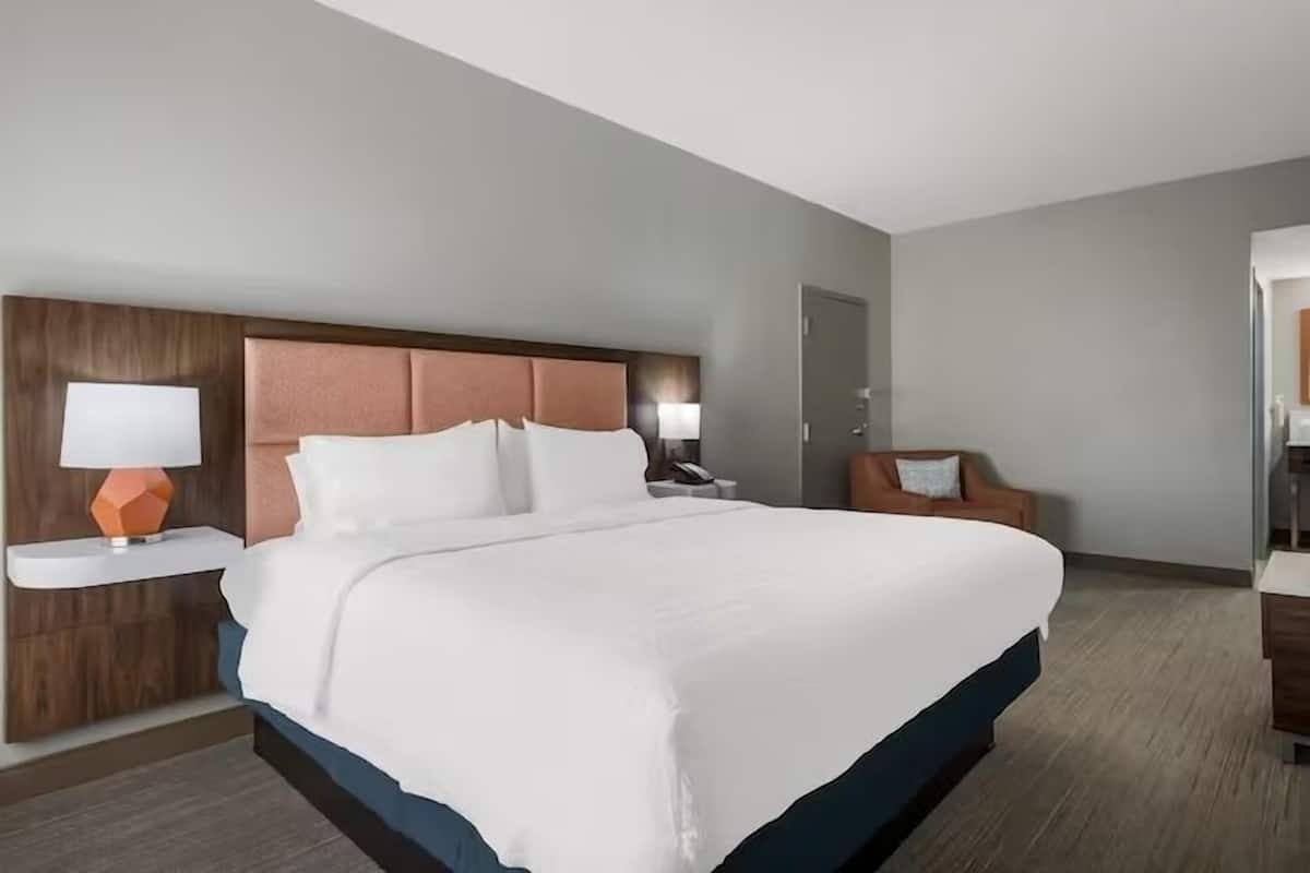 A hotel room with a large bed, a wooden headboard, a nightstand with a lamp, a chair, and a door.