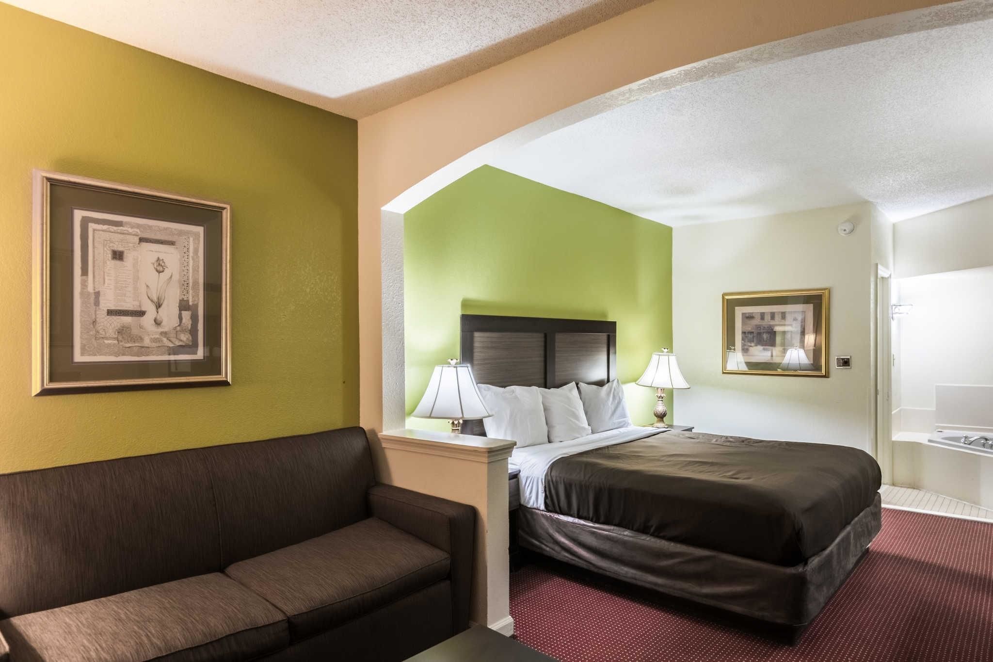A hotel room with a bed, a sofa, a framed picture on the wall, and a bathroom visible through an archway.