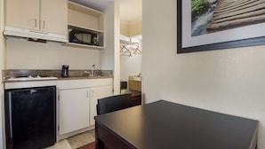 A compact kitchen with a refrigerator, microwave, and sink. There is a dining table with chairs and a small cabinet. A framed picture hangs on the wall. - Suburban Studios Florence I-95 (Florence)