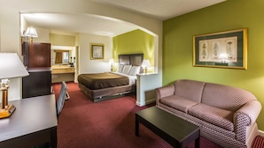 A hotel room with a bed, a sofa, a desk with a lamp, a mirror, and a framed botanical artwork on the wall. - Suburban Studios Florence I-95 (Florence)